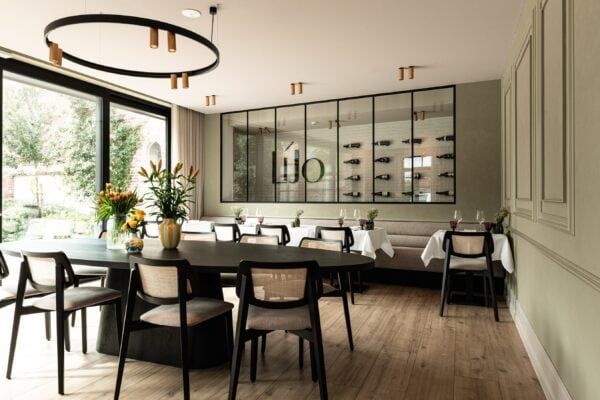 Horecazaal met lange banken op maat, afgewerkt in zachte beige bekleding met fijne stiknaden. Ronde en vierkante tafels met zwarte poten en stoelen in zwart met rotan details zorgen voor een warme, huiselijke sfeer.