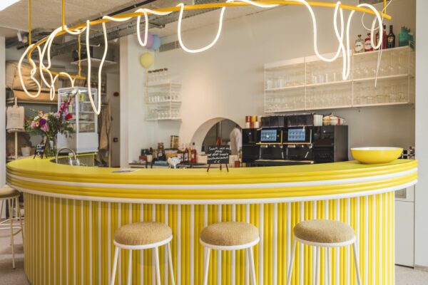 Binnenaanzicht van de barzone met witgelakte Zoa barkrukken en zachte gele stoffering, tegenover een opvallende gele bar in ribstructuur. Voor de bar staan Tina stoelen met houten rug. Dit is bar inrichting horeca met een uitgesproken en uniek kleurpalet.