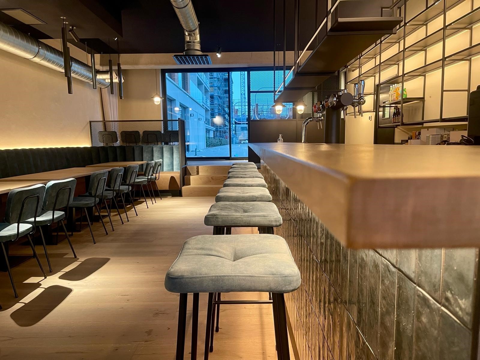 Detail van de barhoek in De Ooverkant Oostende met centraal zicht op een op maat gemaakte horeca bank in zachte fluweelbekleding met diep verticale gebombeerde naden, gecombineerd met een massieve horeca tafel in eik op poottype 6094. De warme houttint van het tafelblad contrasteert mooi met de donkere stoffering en de industriële elementen in het interieur.