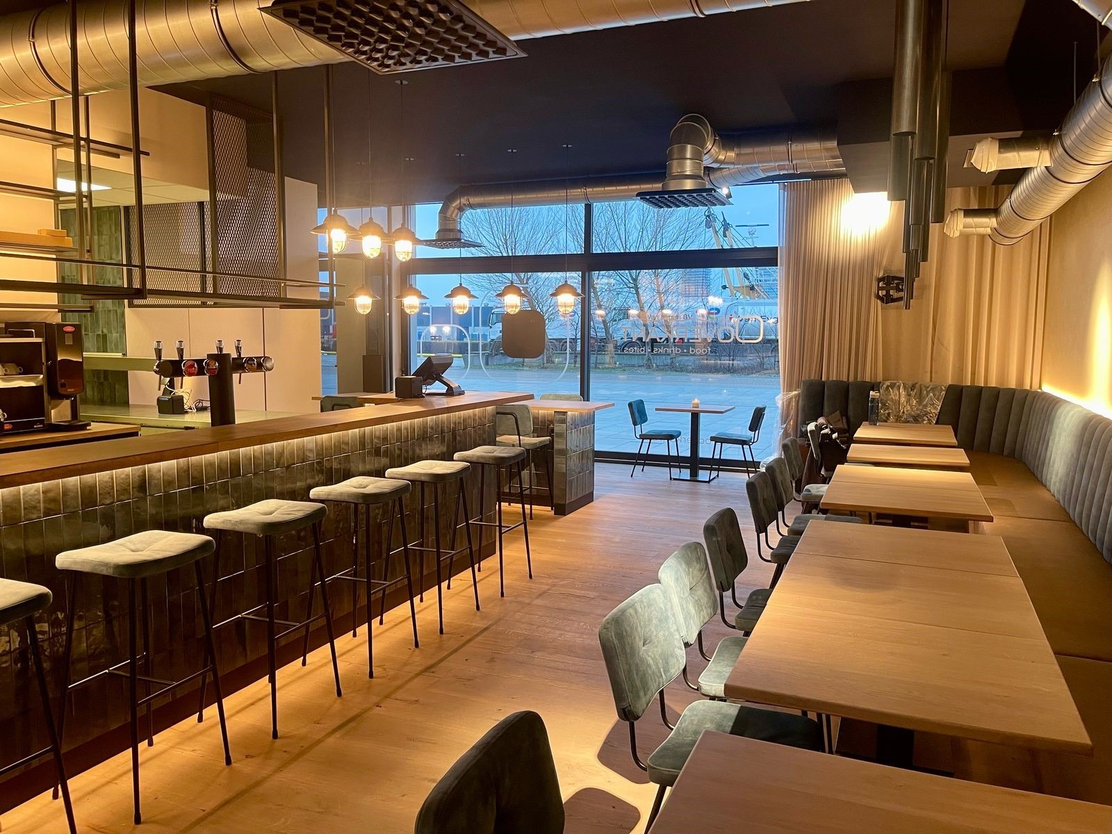 Overzicht van de volledige horecazaak met in de kijker een lange rij maatwerk horeca banken langs de wand en meerdere rechthoekige massief eiken horecatafels op stevige zwarte poten type 6094. De natuurlijke houtstructuur van de tafels versterkt het warme, moderne karakter van de zaak. Perfect voorbeeld van een stijlvolle inrichting horecazaak.