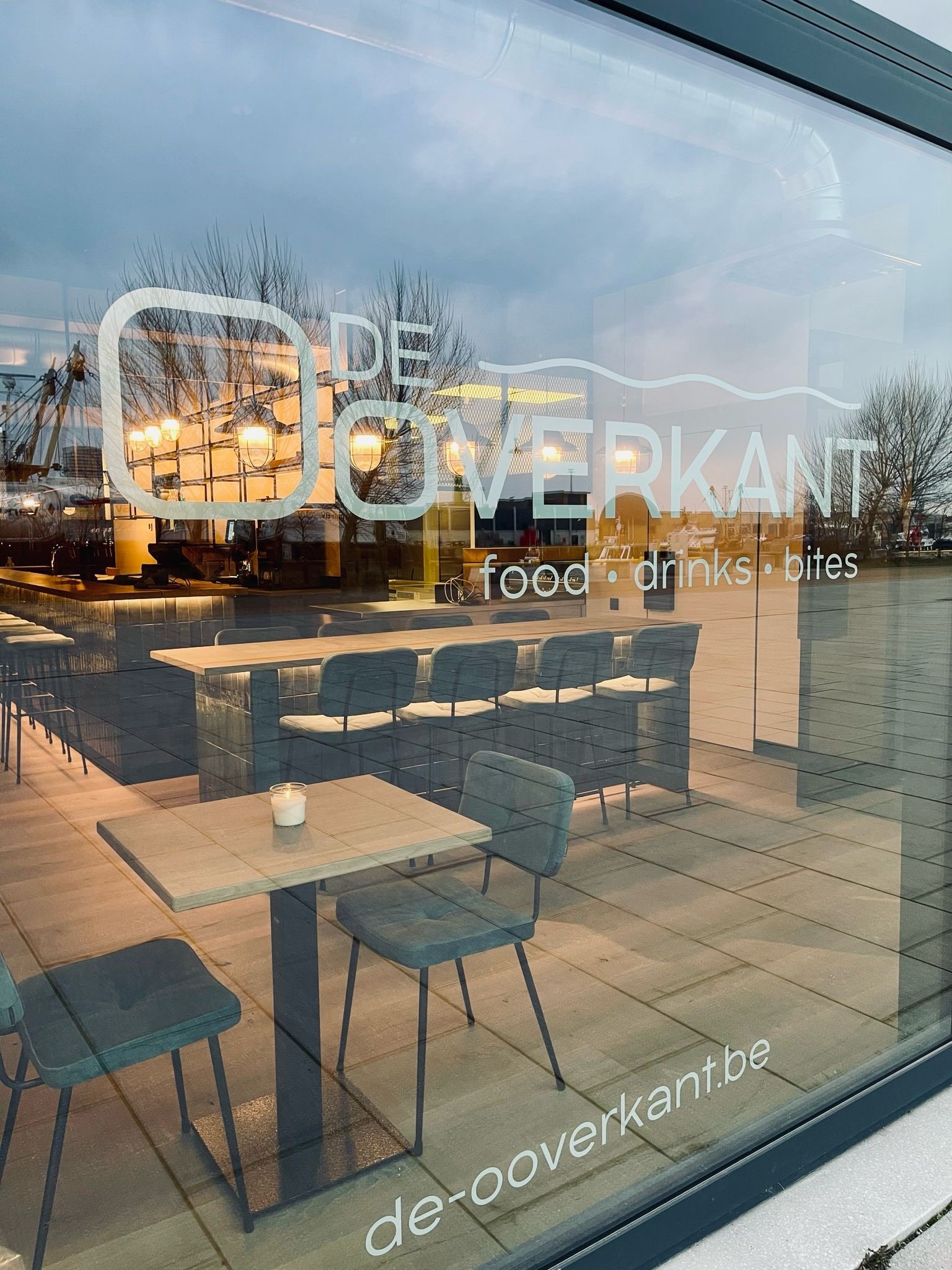 Buitenzicht op De Ooverkant in Oostende met een inkijk naar het interieur waar Búni’s tijdloze horeca tafels in massieve eik duidelijk zichtbaar zijn. De strakke belijning van het eiken blad en het minimalistische poottype 6094 zorgen voor een moderne toets in deze doordachte horeca interieur groothandel-realisatie.