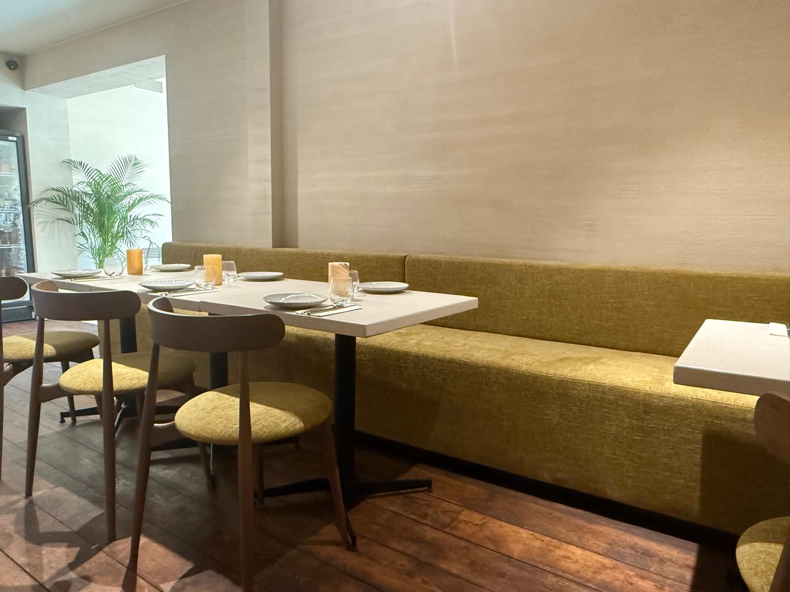 Langgerekte zitopstelling met op maat gemaakte rechte horeca zitbank in fluweelzachte mosterdstof, gecombineerd met witte tafelbladen en houten horeca stoelen met beklede zitting. Deze opstelling garandeert comfort én flexibiliteit in de inrichting horecazaak.