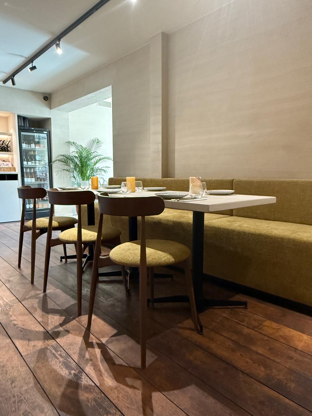 Gezellige tafelhoek in Bistrobar Triple met een combinatie van een rechte zitbank op maat in zachte mosterdgele stof, en houten horeca stoelen met gestoffeerde zit. De contrasterende donkere tafelpoten en het warme parket versterken het stijlvolle karakter van deze horeca interieur inrichting.