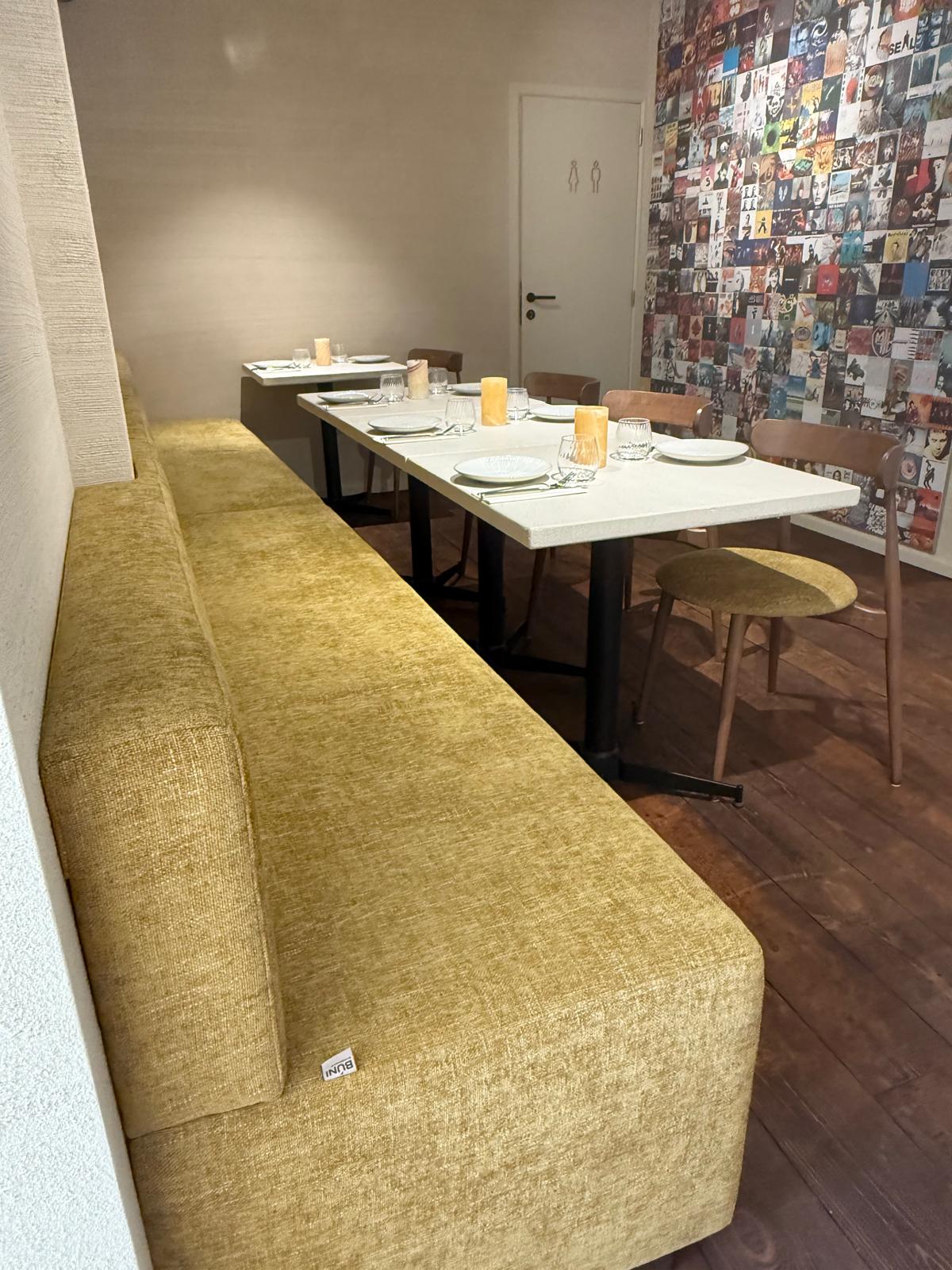 Strakke lijnopstelling met een lange, mosterdkleurige horeca bank op maat in fluwelen stof, gecombineerd met witte tafelbladen en houten stoelen horeca. Deze ruimte ademt comfort en functionaliteit, ideaal voor groepsbezoeken of rustige diners binnen een stijlvol horeca interieur.