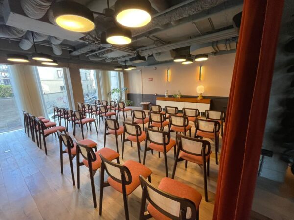 Zicht op een intieme eventruimte in Hotel The Hoxton Brussel met stijlvolle horeca stoelen hout uit de Bilba-reeks. De stoelen hebben een warm notenhouten frame, een beklede zitting in roestkleurige stof en een open rug met fijne gekandeerde rugleuning. De natuurlijke materialen zorgen voor een zachte, akoestisch dempende uitstraling, perfect voor elke horeca interieur inrichting.