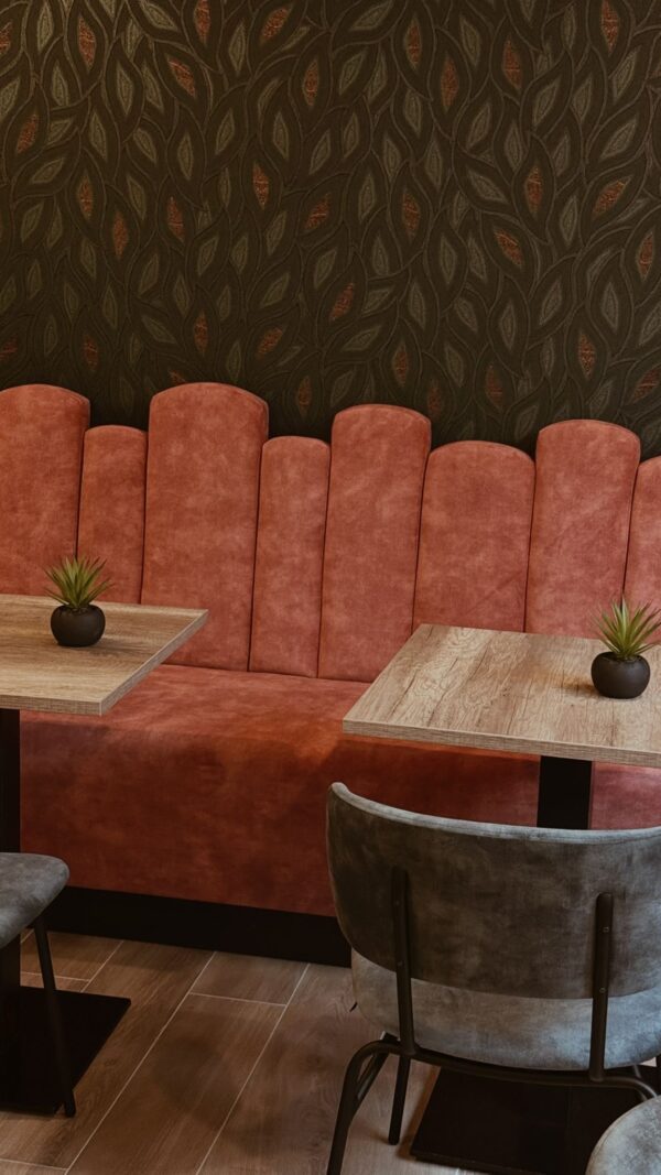 Nabijbeeld van de elegante maatwerk zitbank in Le Champlain. De bank is volledig gestoffeerd in een warme roesttint, met een golvende rug in boogvormen voor extra flair. In beeld ook een deel van de houten horeca tafel met HPL tafelblad en een comfortabele horeca stoel zwart met velours bekleding. Deze combinatie staat garant voor een uitnodigende café inrichting die zowel stijlvol als praktisch is.
