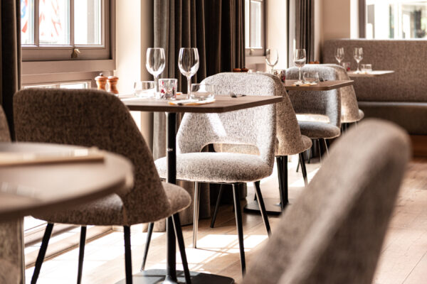 Detailbeeld van een sfeervolle tafelopstelling met comfortabele zitbank op maat langs de wand, aangevuld met stijlvolle Alti-stoelen in beige stof. De horeca tafel met rond HPL-blad en zwart onderstel brengt contrast in het interieur. Perfecte combinatie voor een stijlvolle inrichting restaurant. In samenwerking met Inside – copyright Zeger Vanhaelemeesch