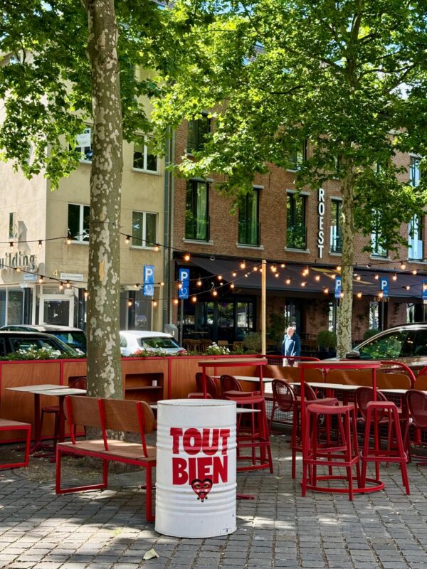 Open terrasopstelling met stapelbare barkrukken horeca in rood gelakt metaal, witte ton-tafels en houten zitbanken met stalen frame. Deze duurzame materialen vormen samen een frisse en uitnodigende horeca terras inrichting.