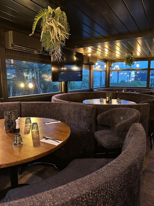 Ruime ronde zitopstelling met een centrale houten tafel en luxueuze Splash horeca stoelen zwart in dezelfde warme bekledingsstof als de maatwerkbanken. De elegante afwerking en natuurlijke accenten maken dit een uitnodigend hoekje binnen een hedendaagse inrichting restaurant.