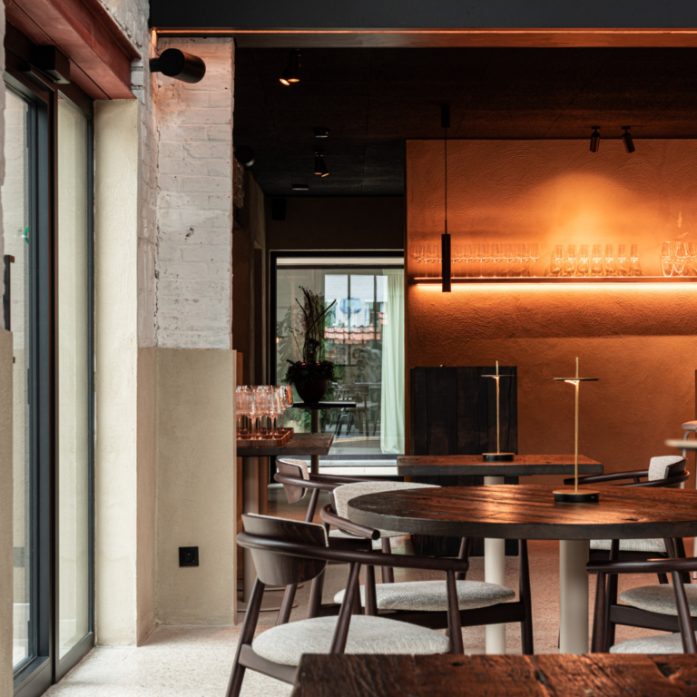 Ronde tafel met vier houten stoelen naast een glazen deur, bakstenen muren en zachte lichtinval via hoge ramen.
