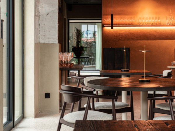 Ronde tafel met vier houten stoelen naast een glazen deur, bakstenen muren en zachte lichtinval via hoge ramen.