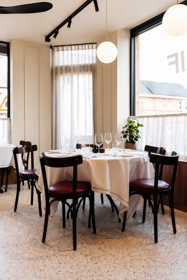 Gezellig ingerichte restauranttafel bij groot raam met witte tafellakens, donkere houten stoelen en warme verlichting.